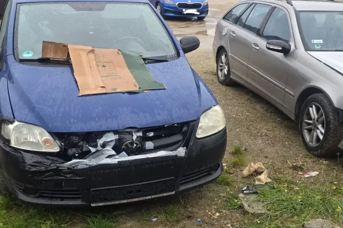 Ein blauer VW mit stark beschädigter, mit Klebeband notdürftig geflickter Frontstoßstange und einem Stück Pappkarton auf der Motorhaube steht auf dem Schotterplatz in der Christianenstraße neben weiteren abgestellten Autos