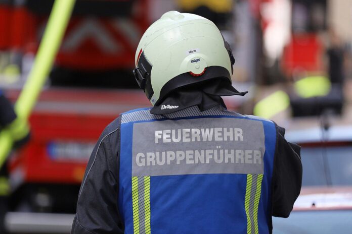 Symbolbild Feuerwehreinsatz Feuerwehrmann mit Schutzhelm und Warnweste mit der Aufschrift Feuerwehr Gruppenführer von hinten vor Einsatzfahrzeugen, Symbolbild
